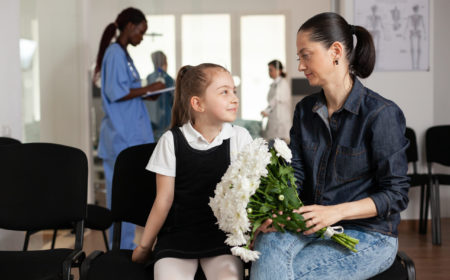 Cheerful family waiting in hospital hallway area during medical examination while black nurse checking clinical insurance documents in background. Mother bringing flowers to sick patient